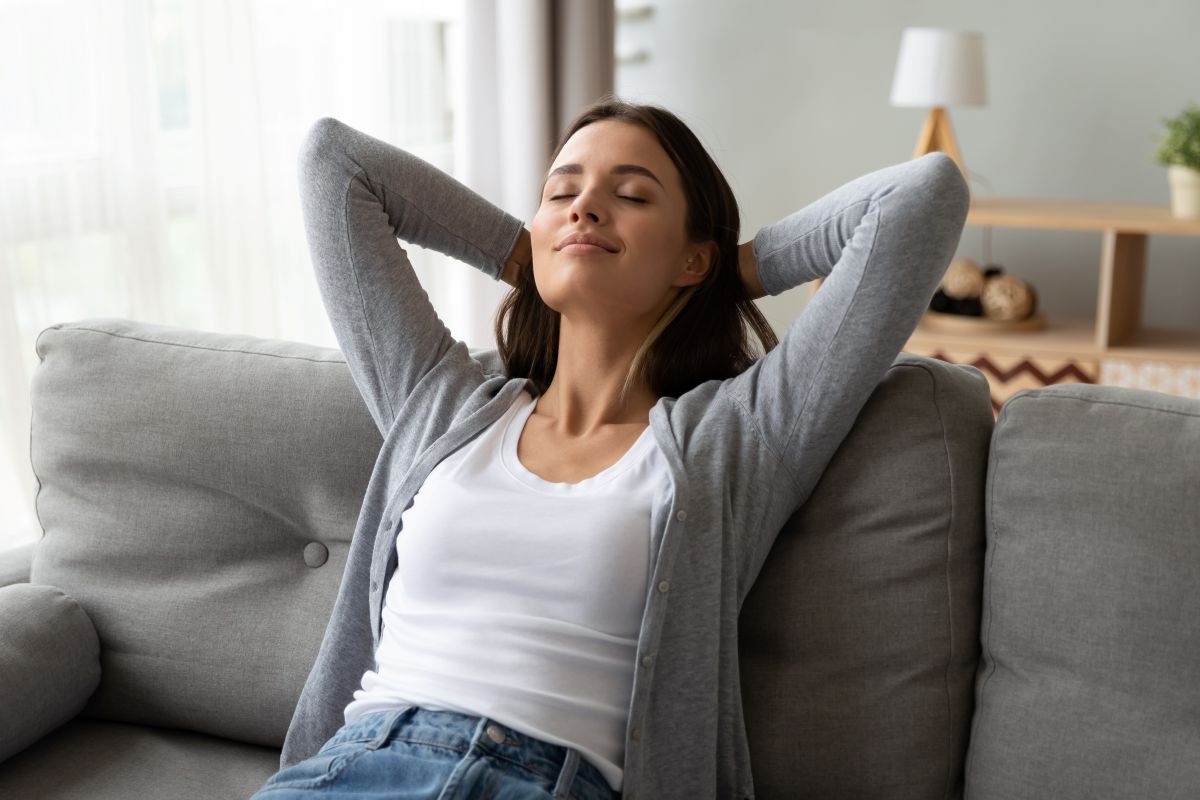 Woman relaxing on a sofa with eyes closed and hands behind head – peaceful home wellness moment